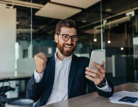 Overjoyed businessman in suit looking at smartphone and clenching fists, making victory gesture in office interior, copy space. Successful business, online win and great deal remote